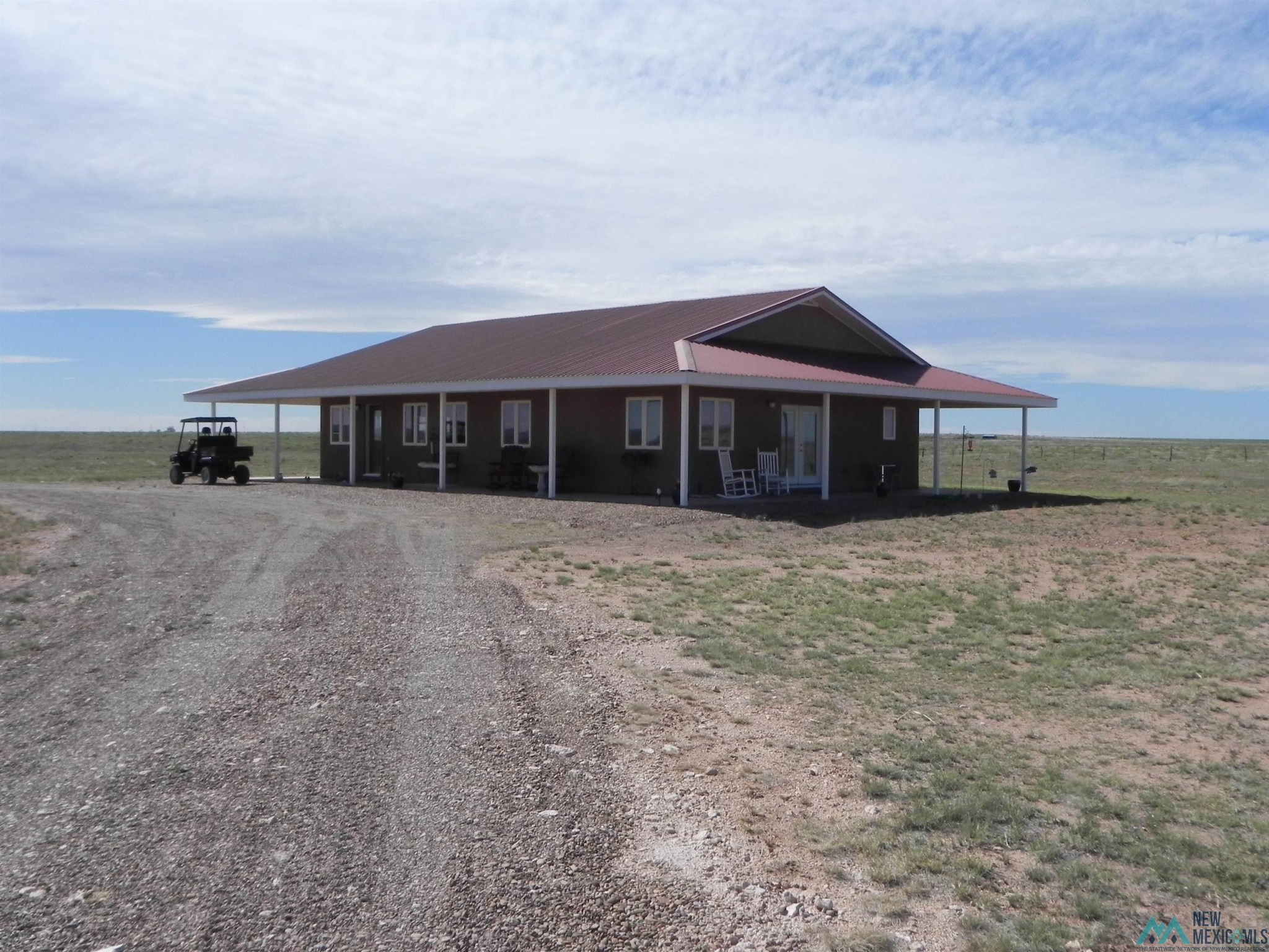 Rural house with a broad metal roof, covered porch, gravel driveway, and utility vehicle on open land.