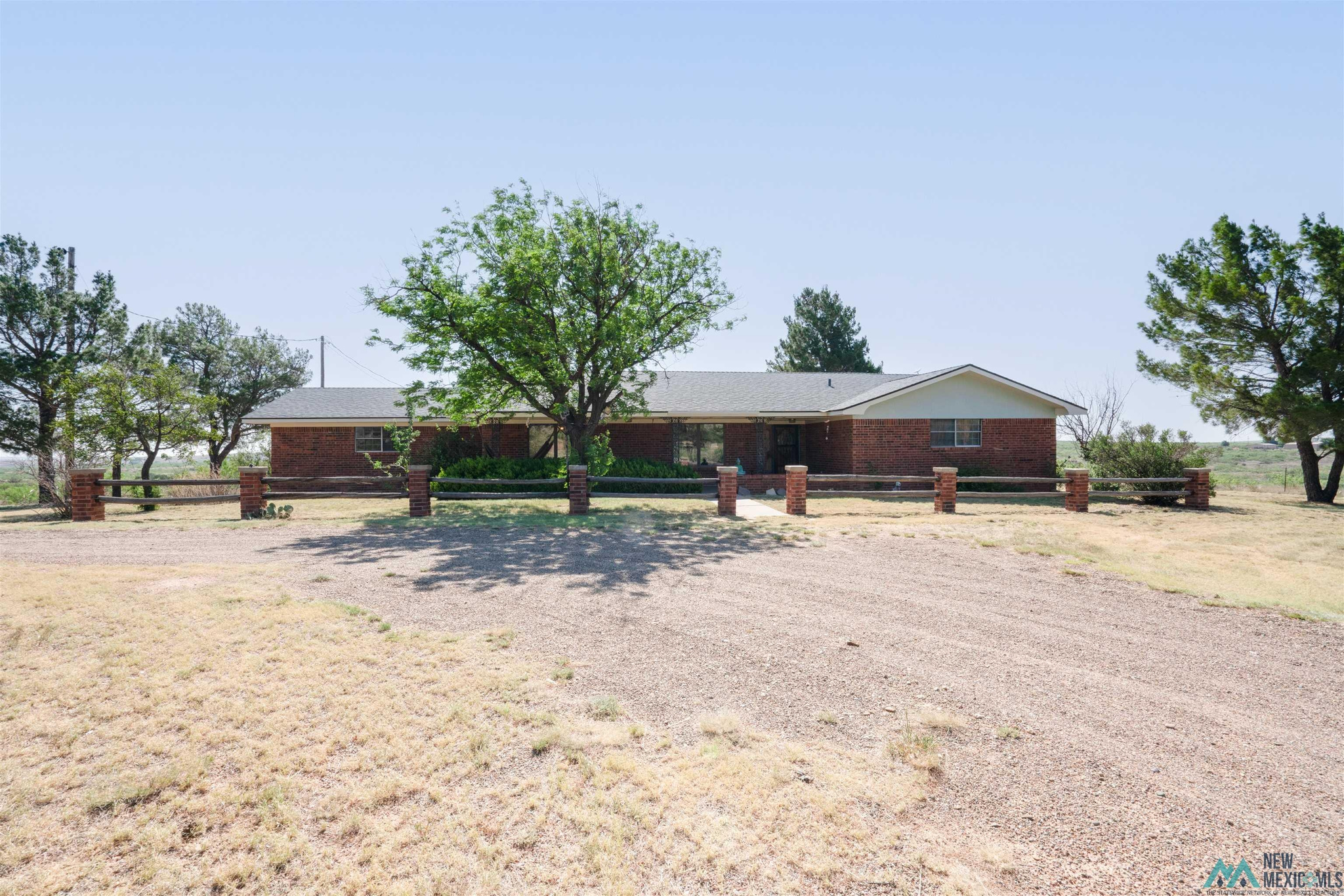 Single-story red brick house behind a low fence, with mature trees and a broad gravel driveway.